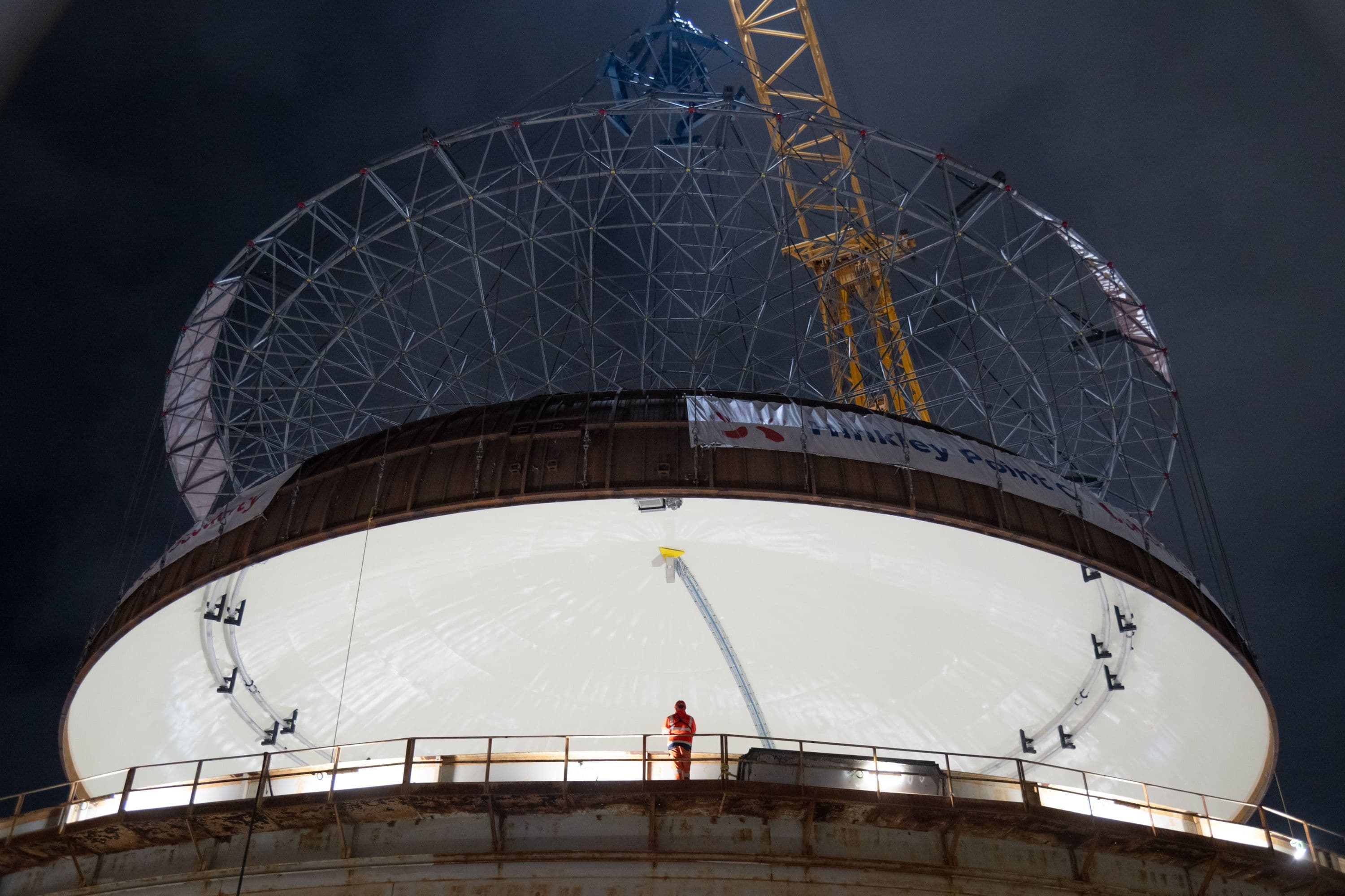 Worker in high-visibility clothing standing on a platform beneath a large circular industrial structure with a lattice framework and crane at night