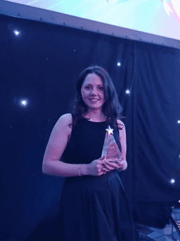 Woman in a black sleeveless dress smiling and holding a star-shaped award on a stage with a dark, star-lit backdrop