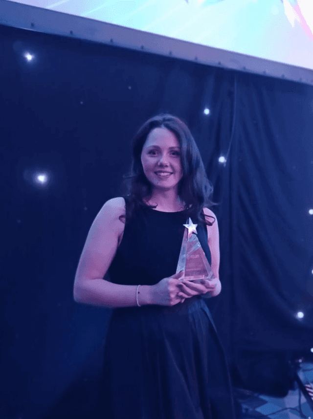 Woman in a black sleeveless dress smiling and holding a star-shaped award on a stage with a dark, star-lit backdrop