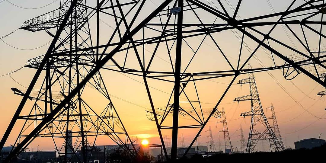 Network of tall electricity pylons and power lines silhouetted against a sunset sky over an industrial landscape