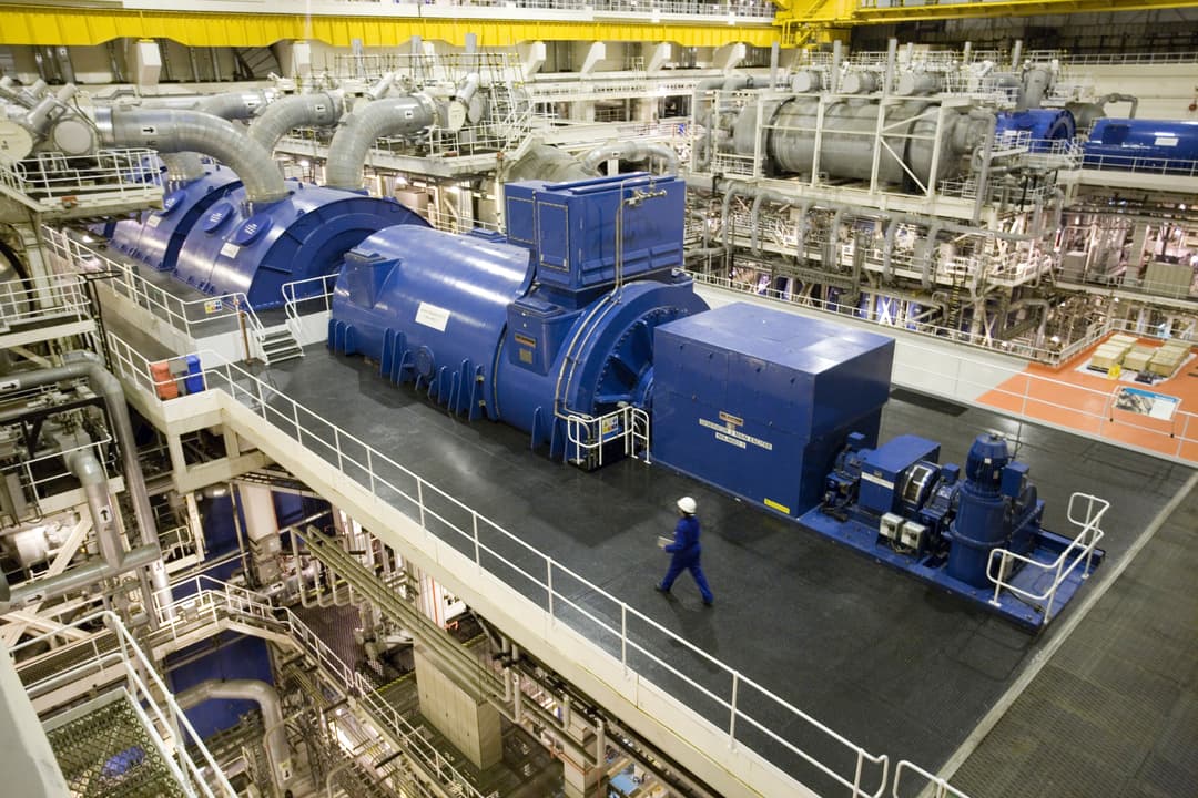Large industrial turbine and generator units inside a power plant with a worker in a hard hat walking along a raised catwalk