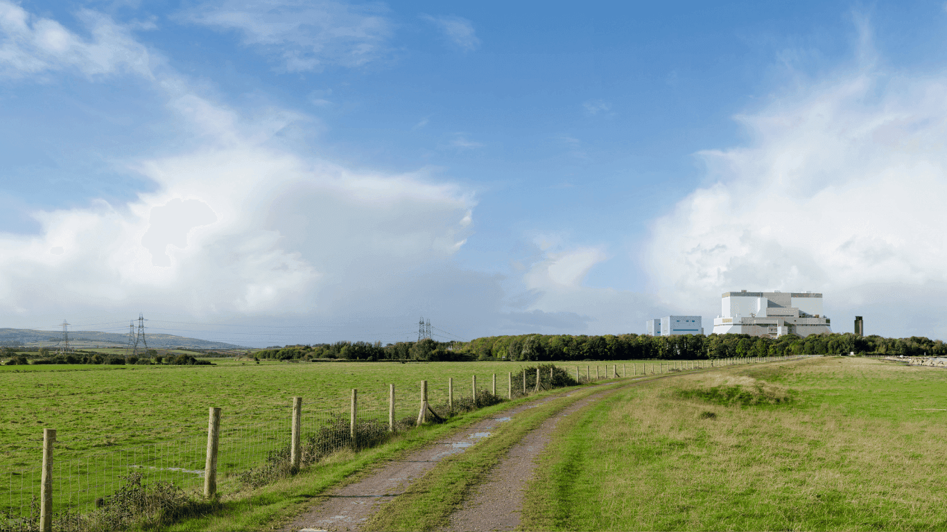 Gravel farm track curving through a green grassy field toward a distant industrial building under a partly cloudy blue sky