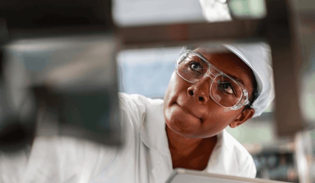 Person in a white hard hat and safety glasses inspecting machinery up close.