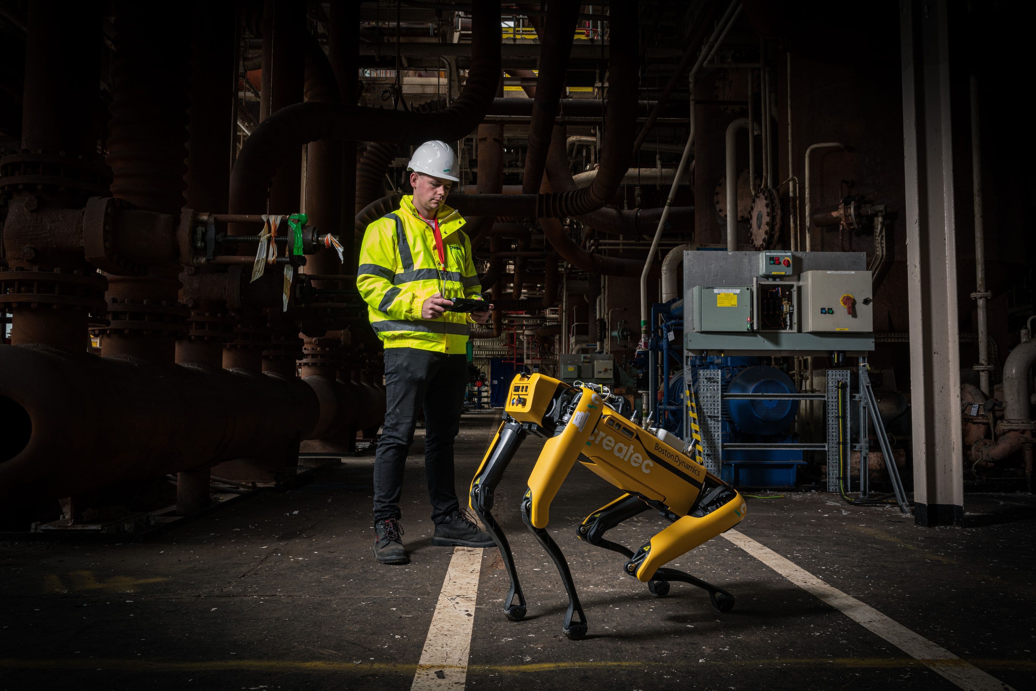 Worker in a high-visibility jacket and hard hat operating a handheld device beside a yellow four-legged robot inside an industrial facility