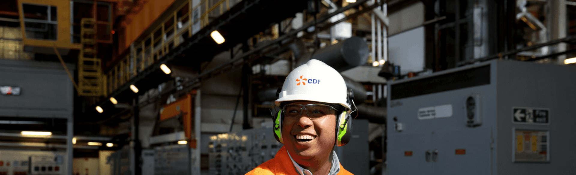 A person wearing a white hard hat with the EDF logo, safety glasses, ear protection, and an orange safety vest smiles inside an industrial facility. The background contains industrial equipment and structures. A person wearing a white hard hat with the EDF logo, safety glasses, ear protection, and an orange safety vest smiles inside an industrial facility. The background contains industrial equipment and structures.