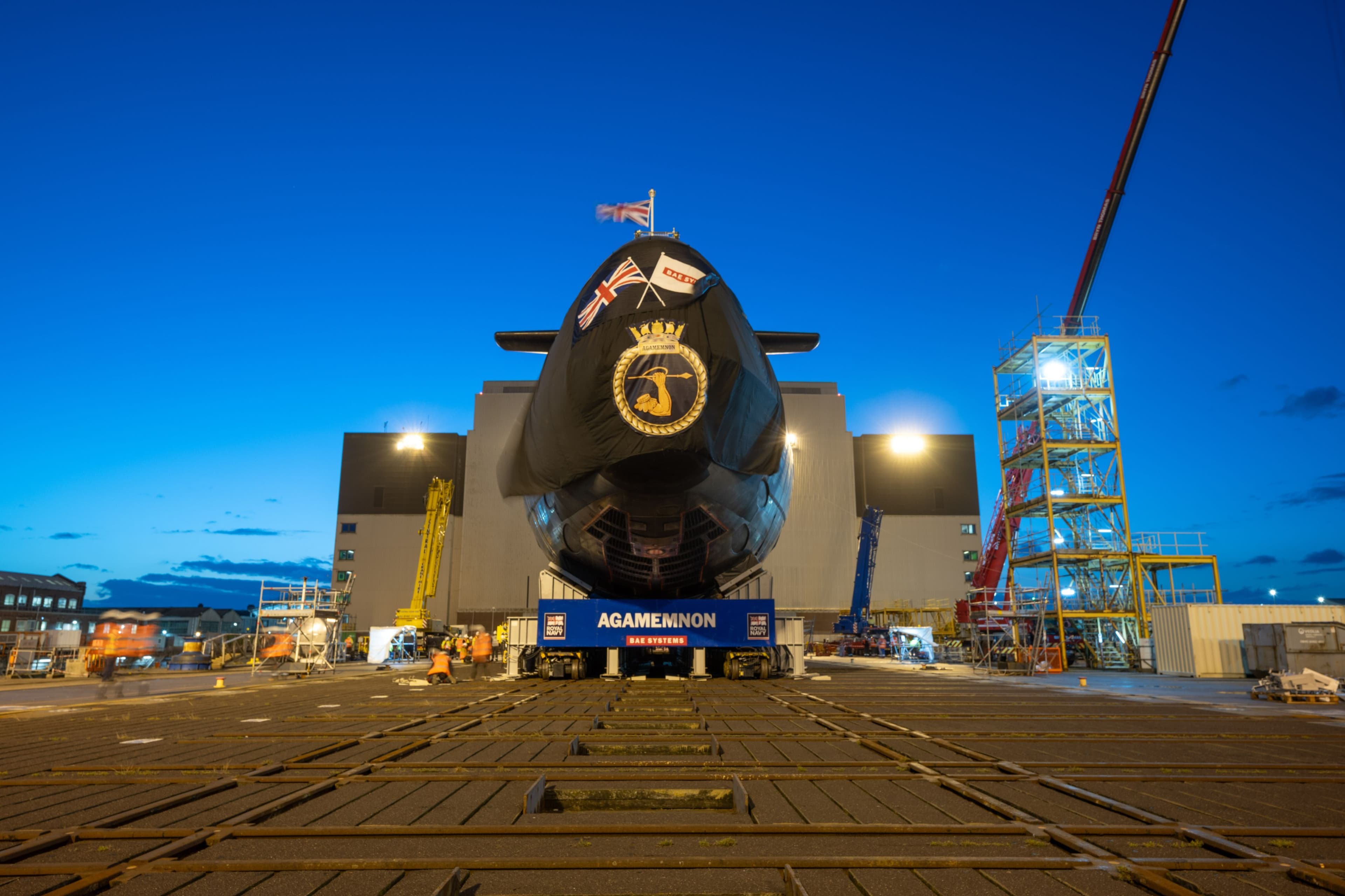 Front view of a submarine on rail tracks in a dockyard at dusk, with cranes, scaffolding, and workers in high-visibility clothing around it