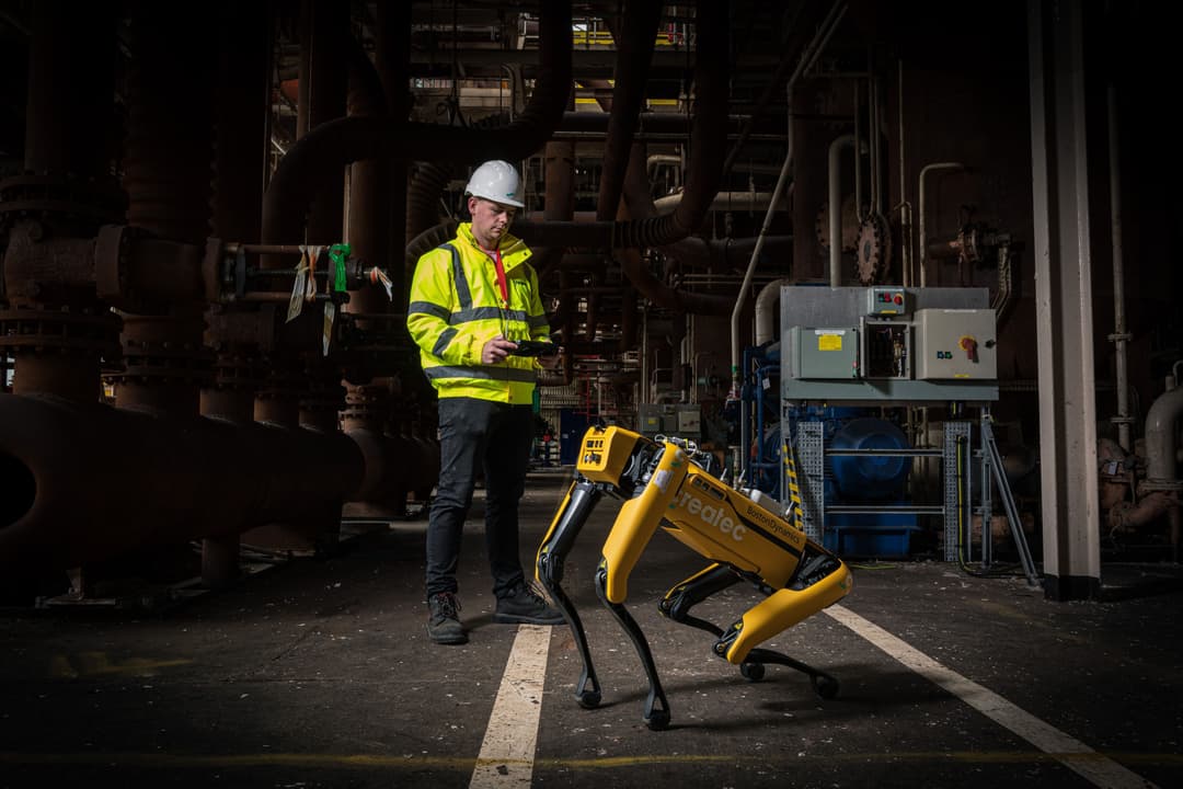 Worker in a high-visibility jacket and hard hat operating a handheld device beside a yellow four-legged robot inside an industrial facility