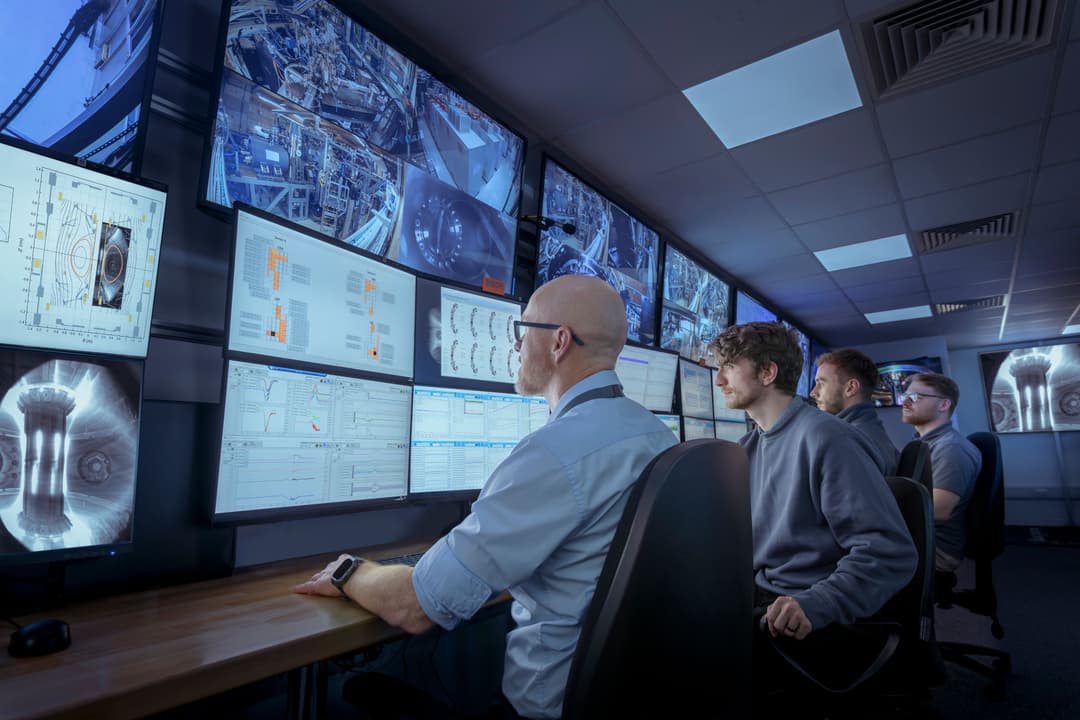 Four workers grouped around multiple screens showing charts and the interior of a nuclear site