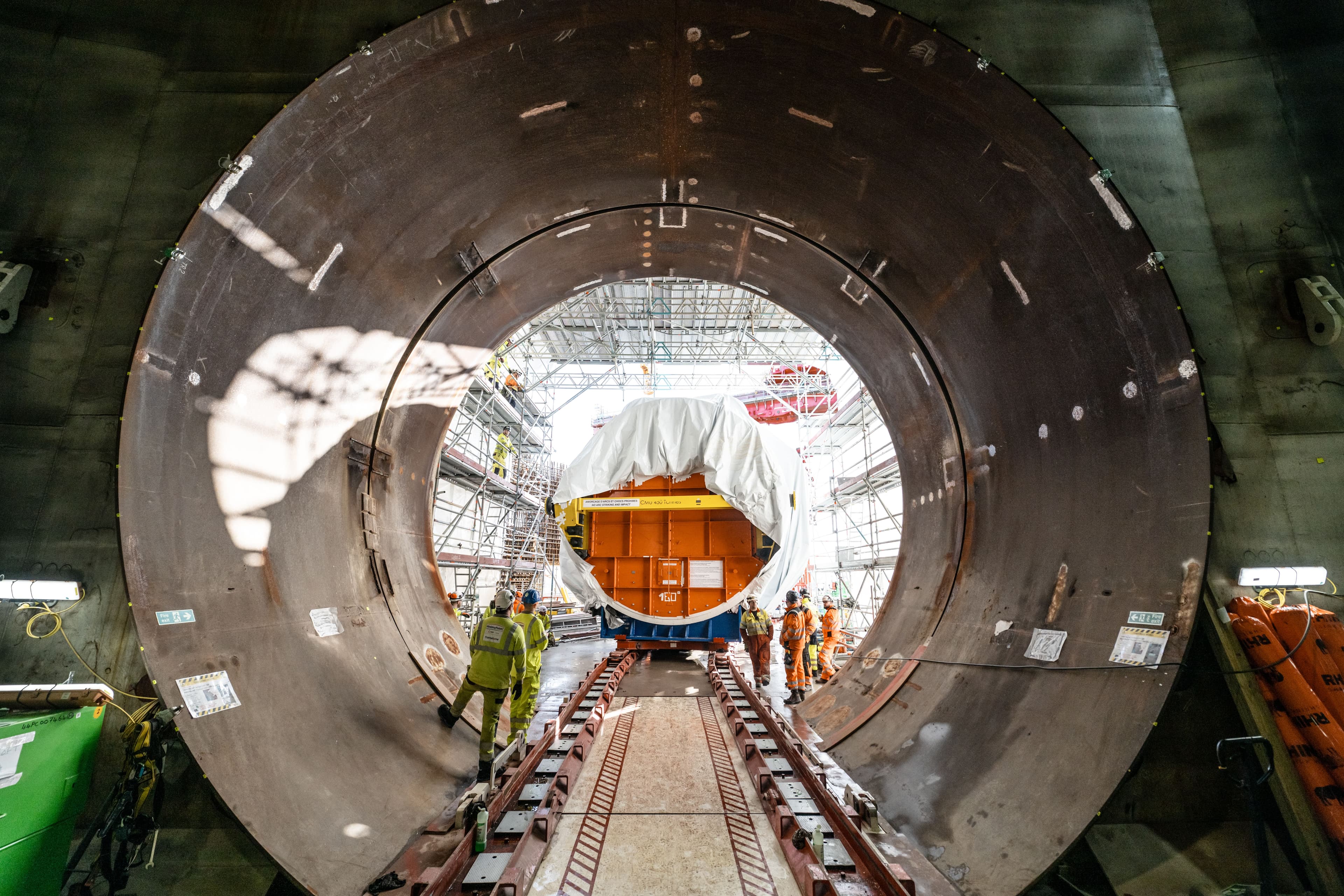 View through a large circular metal tunnel toward a group of workers guiding a covered industrial component along rails inside a construction facility