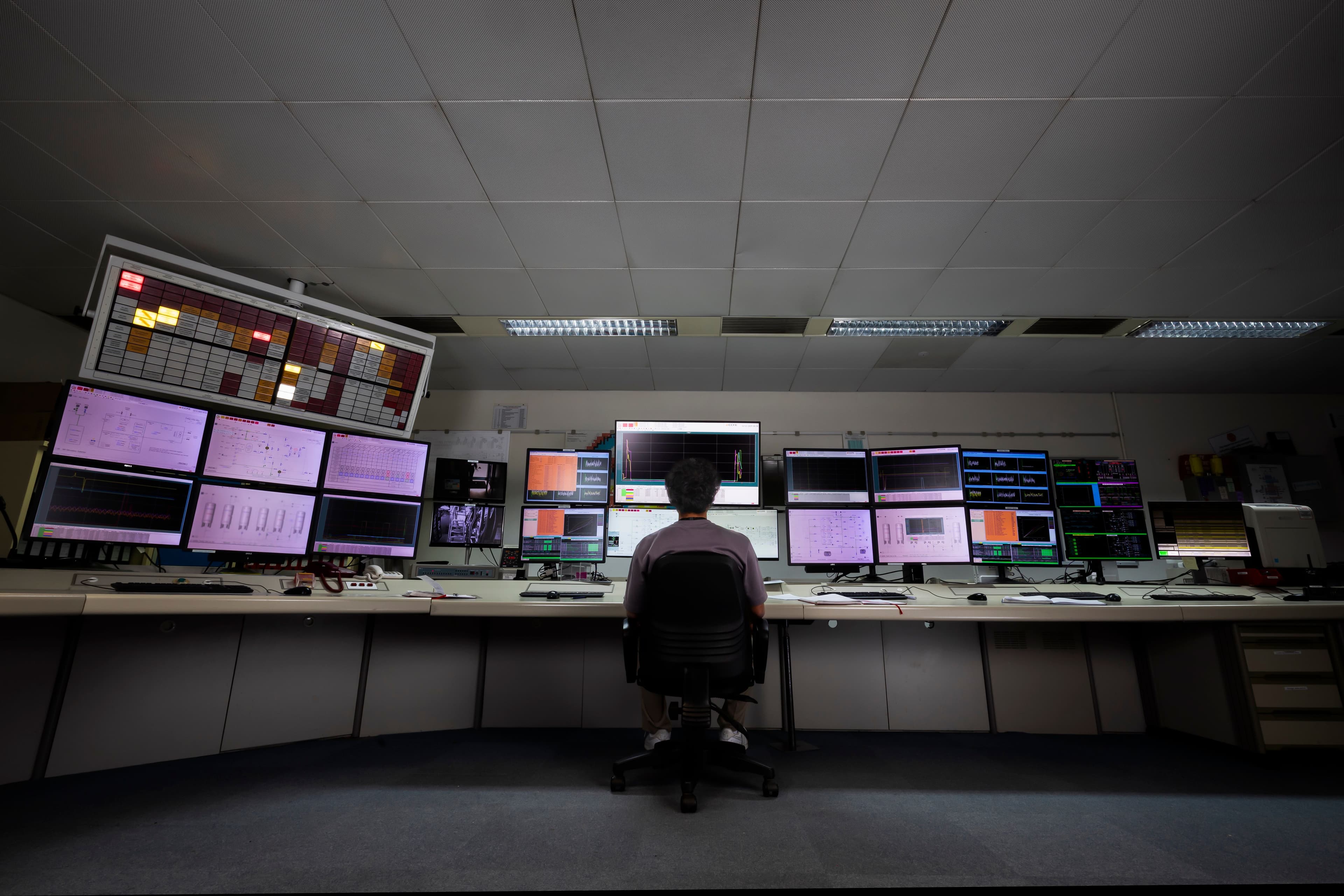 Person seated at a long control desk facing multiple large screens showing technical data and charts.