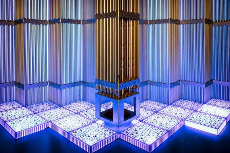 Gold-coloured vertical metal assembly standing on a support frame inside a blue-lit industrial facility with illuminated grid flooring