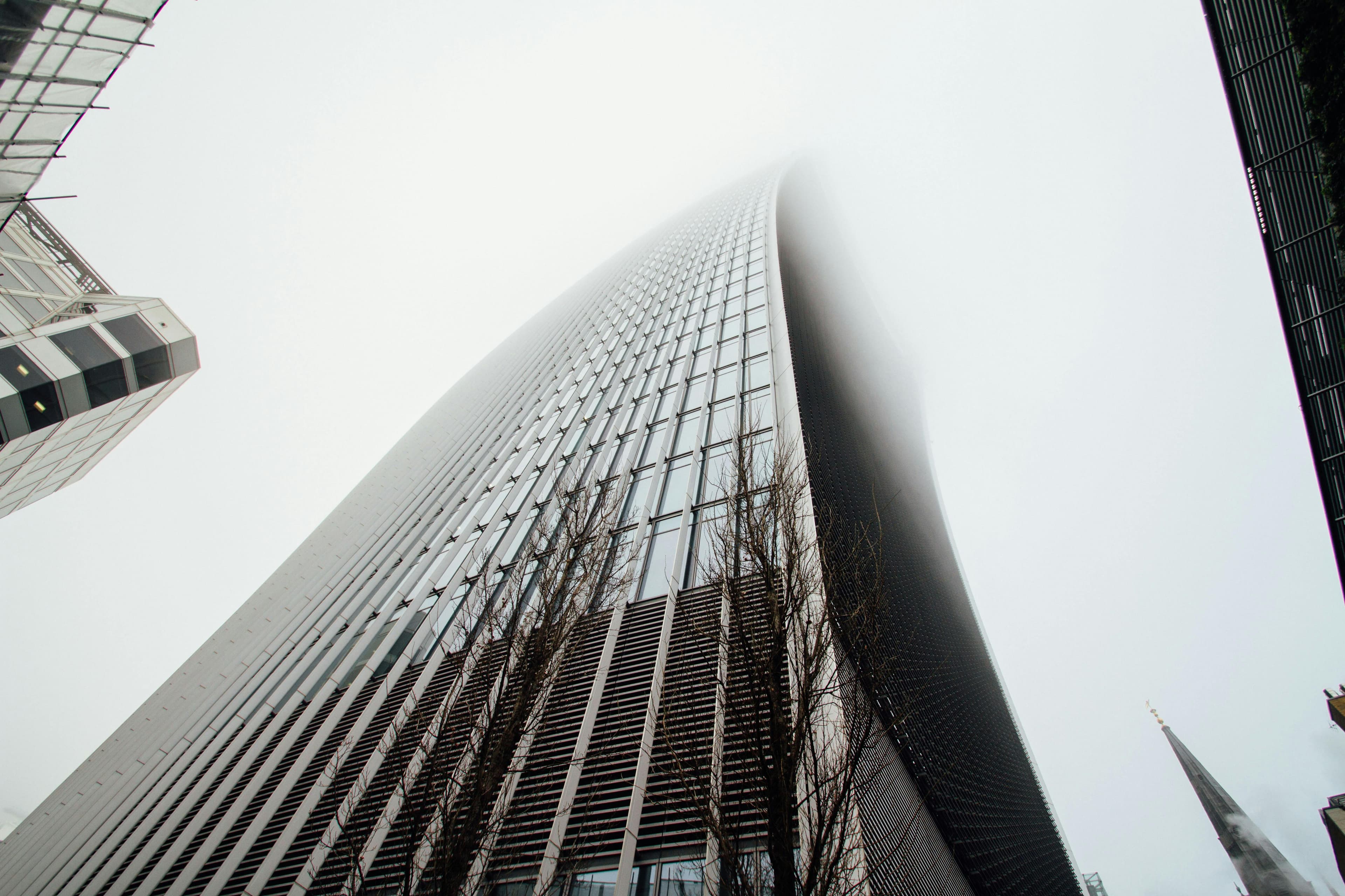 Low-angle view of a modern glass and steel office tower with a curved façade rising into a cloudy sky, flanked by other buildings and bare tree branches at the base.